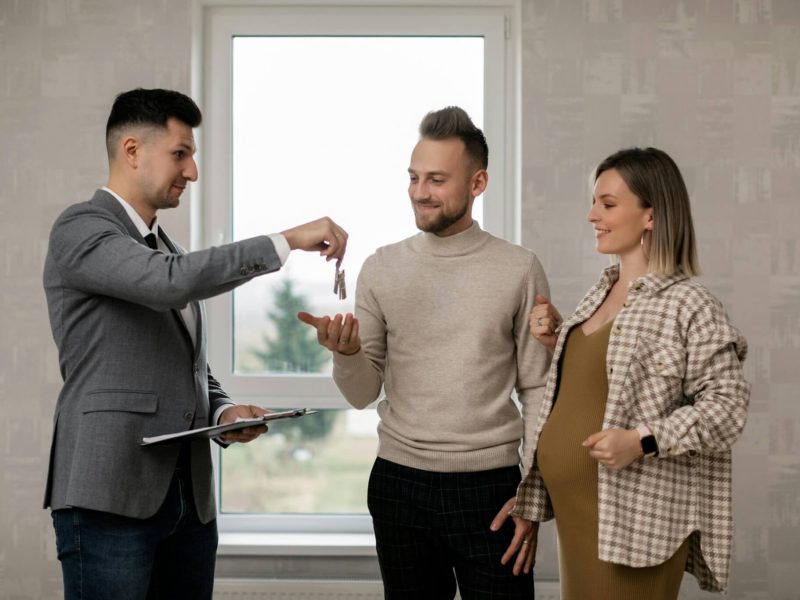 A realtor in a suit dangling keys to hand to a couple, the woman is pregnant